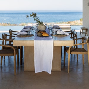 Op de afbeelding: Een wit linnen tafelkleed bedekt een rechthoekige houten tafel met zes stoelen. De tafel is gedekt met borden, glazen en bestek. Een kom met olijven en een brood liggen op de tafel. De tafel is gedekt tegen een achtergrond van een blauwe oceaan en een heldere lucht.