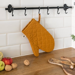 Può includere: Un guanto da forno trapuntato color giallo senape appeso a una cremagliera in metallo nero con ganci. Il guanto da forno è appeso in una cucina con piastrelle bianche a metro e un piano di lavoro in legno. Ci sono mele e noci sul piano di lavoro.