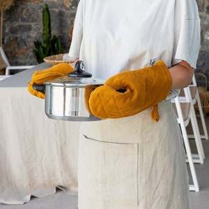 Op de afbeelding: Een persoon draagt een wit shirt en een linnen schort en houdt een roestvrijstalen pan met deksel vast met behulp van twee gele, gewatteerde ovenwanten. Het schort heeft een grote zak aan de voorkant. De tekst "THE LINEN VALLEY" is zichtbaar aan de rechterkant van de afbeelding.