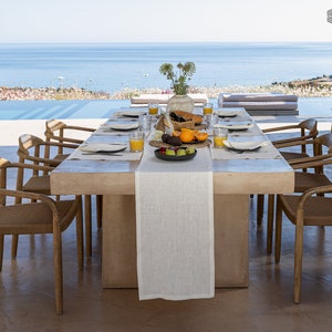 Può includere: Un lungo tavolo rettangolare in legno marrone chiaro con una tovaglia di lino bianco e un runner bianco. Il tavolo è apparecchiato con piatti, bicchieri e posate. Ci sono anche ciotole di frutta e un piatto di cibo sul tavolo. Il tavolo è davanti a una grande finestra con vista sull'oceano. Il testo "THE ZEN VALLEY" è visibile nell'angolo in alto a destra dell'immagine.