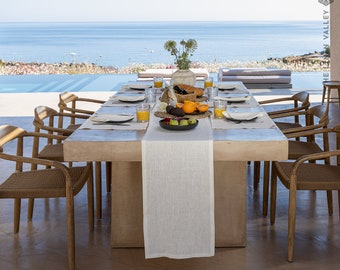 May include: A long, rectangular, light brown wooden table with a white linen tablecloth and a white runner. The table is set with plates, glasses, and cutlery. There are also bowls of fruit and a plate of food on the table. The table is in front of a large window with a view of the sea. The text "THE ZEN VALLEY" is visible in the upper right corner of the image.