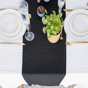 Pode incluir: Um corredor de mesa de linho preto sobre uma toalha de mesa de linho branco. Duas configurações de lugar com pratos brancos e talheres dourados estão na toalha de mesa. Um copo de água e uma tigela pequena de azeitonas estão no corredor de mesa. Uma planta em vaso com folhas verdes está no centro do corredor de mesa.