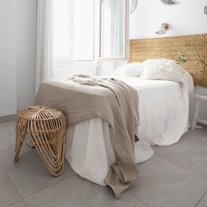 Puede incluir: Una cama blanca con una cabecera tejida y una manta de lana beige. Un taburete tejido se encuentra junto a la cama.