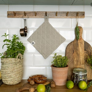 Può includere: Un presina pentola in lino beige è appeso a una mensola di legno con ganci. Il presina pentola ha un design trapuntato. Ci sono vari oggetti da cucina su un bancone sotto la mensola, tra cui un tagliere di legno, un barattolo di vetro e un cestino.