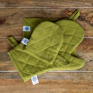 May include: Two green quilted oven mitts and a matching green quilted pot holder. The mitts have a loop handle and the pot holder has a label that says "The Linen Valley".
