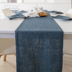 Könnte beinhalten: Ein blauer Tischläufer aus Leinen auf einem weißen Tisch, mit passenden Servietten und Tellern. Ein Brotlaib und eine Tasse stehen auf dem Tisch. Der Läufer hat eine strukturierte Oberfläche.