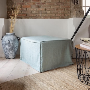 Op de afbeelding: Een grote, vierkante poef bedekt met een lichtblauwe stof. De poef staat op een lichtgekleurde houten vloer, met een geweven vloerkleed ervoor. Een decoratieve vaas met gedroogde takken en een kleine bijzettafel zijn ook zichtbaar.