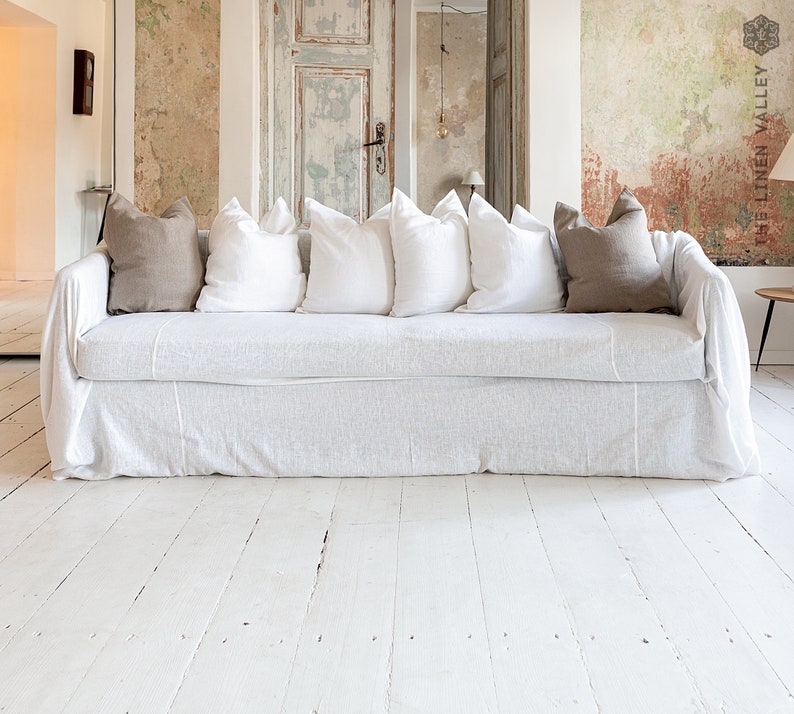 May include: A white slipcovered sofa featuring a long, draped fabric cover. The sofa is accessorized with four white square throw pillows and two taupe square throw pillows arranged along the backrest, creating a layered and textured appearance.