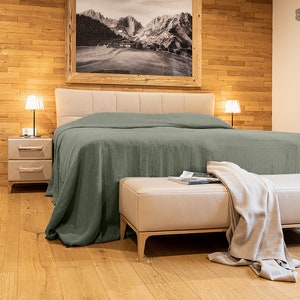 May include: A green linen duvet cover on a bed with a light brown wooden headboard. A white leather ottoman with a book on top sits in front of the bed. The bed is in a room with a wood-panelled wall and a framed black and white photograph of a mountain scene.