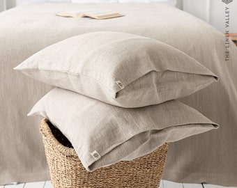 Puede incluir: Dos cojines cuadrados hechos de tela de lino beige claro apilados sobre una cesta de mimbre tejida. Cada cojín presenta una pequeña etiqueta de tela blanca en el lateral con el texto THE LINEN VALLEY impreso en un color oscuro.