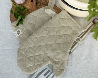 May include: A pair of quilted oven mitts made from natural beige linen fabric. Each mitt features a diamond-stitched pattern, a fabric hanging loop, and a small white rectangular tag with the text The Linen Valley printed in black.