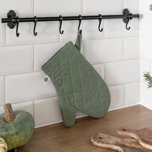 May include: A sage green quilted oven mitt with a fabric loop for hanging. The mitt features a diamond-stitched pattern and a small white brand tag on the side. It is suspended from a black metal hook on a wall-mounted rack above a wooden counter.