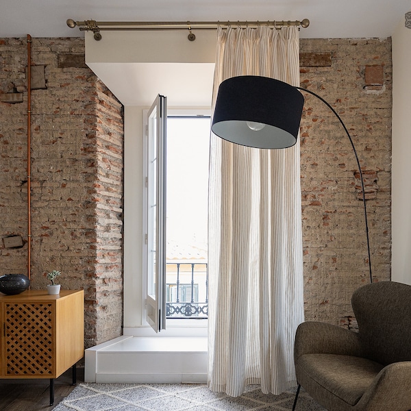 May include: A living room with a large window, white curtains with thin stripes, a brown armchair, and a floor lamp with a black shade. The wall is made of exposed brick.
