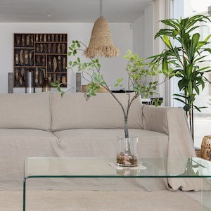 Peut inclure: Un canapé en lin beige clair avec une couverture, une table basse en verre et une plante en pot dans un vase en verre transparent. Le canapé est dans un salon moderne avec un mur blanc et une grande fenêtre.