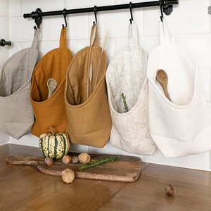 Può includere: Cinque sacchetti portaoggetti in lino appesi a un'asta di metallo nero. I sacchetti sono di vari colori, tra cui beige, giallo senape e bianco. I sacchetti sono riempiti con oggetti come fiori secchi, rametti di rosmarino e cucchiai di legno. I sacchetti sono etichettati "THE LINEN VALLEY".