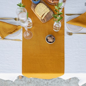 Puede incluir: Un camino de mesa de lino amarillo mostaza sobre un mantel de lino blanco. Hay dos cubiertos con cubiertos de oro, servilletas y vasos. Un pequeño cuenco de nueces, un plato de queso y un jarrón de vegetación también están en la mesa. El texto "THE LINEN VALLEY" es visible en la esquina superior derecha de la imagen.