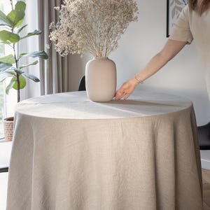 Puede incluir: Un mantel redondo de lino beige cubre una mesa. Un jarrón beige contiene flores secas. La escena está ambientada en una habitación luminosa con una ventana y una planta. Un brazo se extiende hacia la mesa.