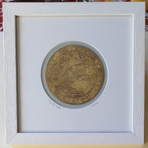 Op de afbeelding: Een gouden metalen plaquette met een gegraveerd beeld van een persoon die een kinderwagen duwt onder een paraplu. De plaquette is ingelijst in een wit frame en heeft de tekst "Double Happy" en "Oliver" erop.