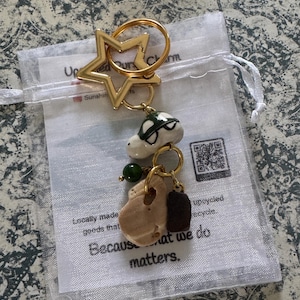 May include: Gold-tone star-shaped keychain with a ring, holding charms including a turtle, a stone, and a dark brown rectangle. The keychain is packaged in a clear bag with text that reads "Because what we do matters."