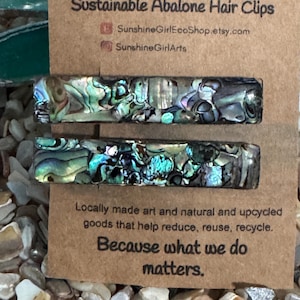 May include: Two iridescent abalone shell hair clips on a brown card with the text "Sustainable Abalone Hair Clips" and "SunshineGirlEcoShop.etsy.com" and "SunshineGirlArts". The card also says "Locally made art and natural and upcycled goods that help reduce, reuse, recycle. Because what we do matters."