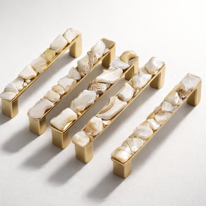 May include: Five decorative cabinet pulls with a rectangular design. Each pull features a gold-colored metal base and is adorned with a mosaic of white and beige stones. The pulls are arranged on a white surface.