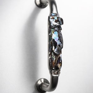 以下が含まれることがあります： アワビの貝殻の装飾が施されたステンレス製のドアプル。ハンドルは湾曲したデザインで、白い表面に取り付けられています。アワビの貝殻は、青、緑、茶色の虹色の色をしています。