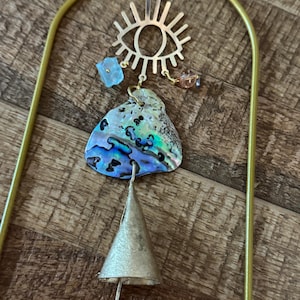 Könnte beinhalten: Goldfarbener Metall-Wandbehang mit einem augenförmigen Anhänger, blauen und braunen Perlen, einer Abalone-Muschel und einer Glocke. Der Wandbehang hängt an einem goldfarbenen Bogen.