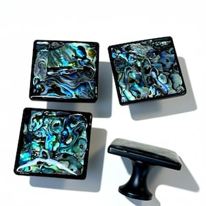 Puede incluir: Cuatro tiradores cuadrados para armarios con incrustaciones iridiscentes de concha de abulón y marcos negros. Un tirador es un asa con base negra. Los tiradores tienen un acabado brillante sobre un fondo blanco.