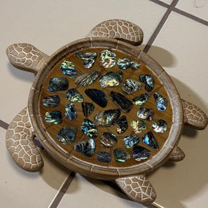 May include: A turtle-shaped decorative object with a light brown, textured wooden frame and flippers. The shell area is filled with a smooth, amber-toned resin inlay embedded with numerous small, iridescent abalone shell pieces in blue, green, and purple hues.