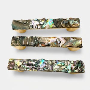 May include: Three rectangular cabinet pulls featuring a gold-toned metal base topped with an inlay of iridescent abalone shell fragments. Each handle displays a unique mosaic of natural shell pieces in shades of green, blue, and grey.