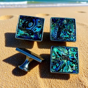 Puede incluir: Cuatro tiradores cuadrados para armarios con incrustaciones de concha de abulón iridiscente. Los tiradores tienen un marco de metal plateado y un vástago a juego. Los tiradores se exhiben sobre una superficie arenosa con el océano al fondo.