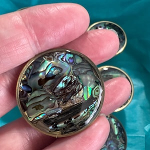 May include: A set of circular buttons featuring a polished abalone shell inlay with iridescent shades of green, blue, and purple, encased in a smooth gold-toned metal rim. The natural shell pattern displays varied textures and dark organic veins.