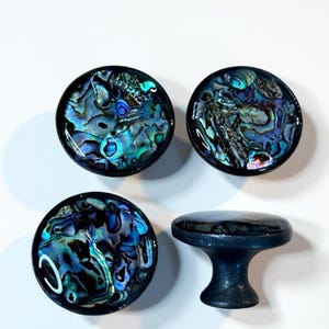 Può includere: Quattro pomelli decorativi per mobili con intarsi di conchiglia di abalone iridescente. I pomelli sono rotondi con un bordo nero e la conchiglia di abalone presenta sfumature di blu, verde e viola. Un pomello è una maniglia con finitura blu scuro.