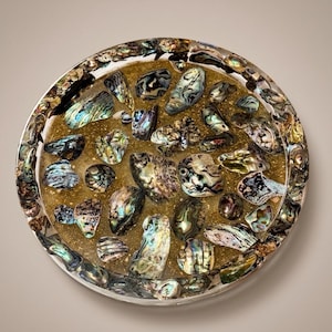 Op de afbeelding: Een ronde onderzetter met een basis van heldere hars, ingelegd met talloze iriserende abalone-schelpfragmenten en fijne gouden glitters. De schelpstukjes vertonen een verscheidenheid aan kleuren, waaronder groen, blauw en paars.