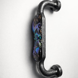 以下が含まれることがあります： C字型のキャビネットプルハンドル。ハンドルは暗い質感の金属製で、虹色のあわびの貝殻の象嵌が施されています。アワビの貝殻の象嵌は、青、緑、紫の色合いを示しています。