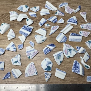 May include: A collection of broken ceramic pieces with blue and white floral and geometric patterns. The fragments vary in size and shape, scattered on a wooden surface. Some pieces have pink floral accents.