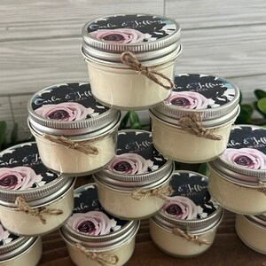 以下が含まれることがあります： 蓋付きの小さなガラス瓶が積み重ねられており、それぞれに白いろうそくが入っています。瓶は、花柄と「Carla & Jeffrey 08.19.23」の文字が書かれた白黒のラベルで飾られています。