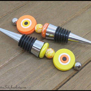 May include: Two decorative bottle stoppers with a silver metal base and a black rubber stopper. Each stopper is topped with a colorful eye-shaped bead with a white, orange, and black design. The beads are connected to the stopper with a string of small white and orange beads.