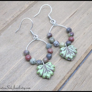 May include: A pair of silver hoop earrings with green leaf charms. The hoops are adorned with multi-colored beads, including red, brown, and green.