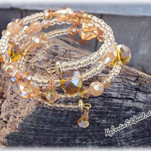 Puede incluir: Pulsera envolvente de m&uacute;ltiples hilos con una mezcla de cuentas y cristales dorados y &aacute;mbar. La pulsera tiene un dise&ntilde;o en espiral con dijes colgantes. Se exhibe sobre un trozo de madera envejecida.