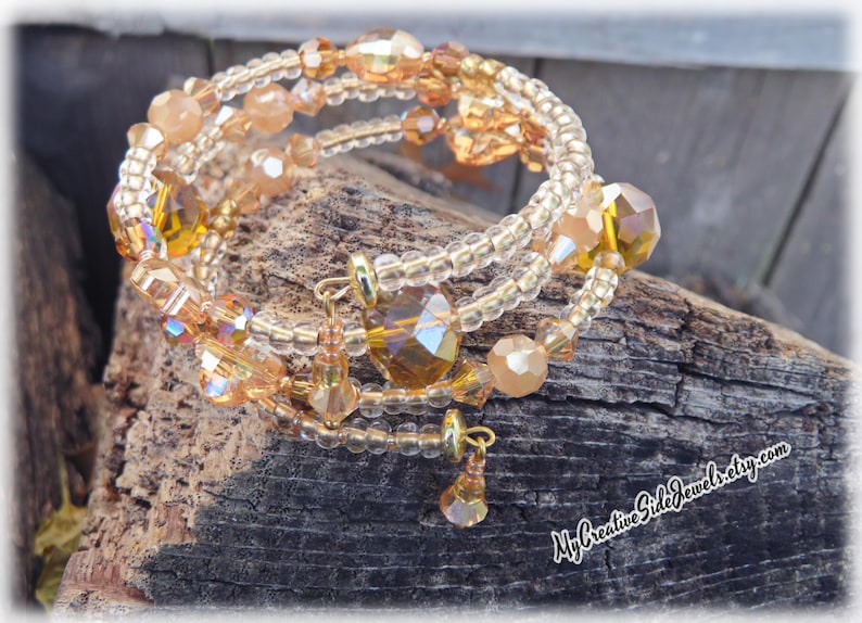 Puede incluir: Pulsera envolvente de m&uacute;ltiples hilos con una mezcla de cuentas y cristales dorados y &aacute;mbar. La pulsera tiene un dise&ntilde;o en espiral con dijes colgantes. Se exhibe sobre un trozo de madera envejecida.