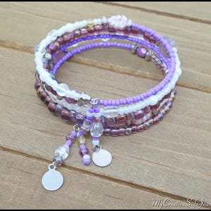 May include: A multi-strand bracelet with purple, white, and burgundy beads. The bracelet features a spiral design with dangling charms and small silver discs. The beads are arranged in layers, creating a textured and colorful accessory.