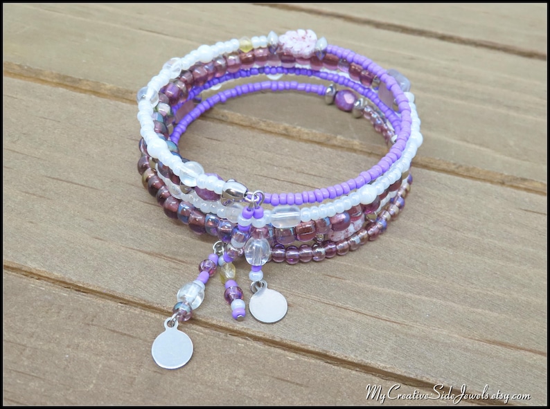 May include: A multi-strand bracelet with purple, white, and burgundy beads. The bracelet features a spiral design with dangling charms and small silver discs. The beads are arranged in layers, creating a textured and colorful accessory.