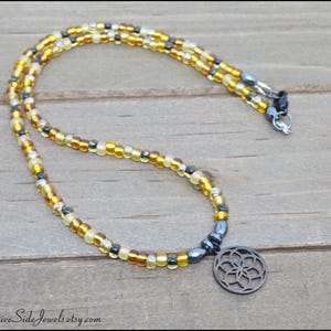 May include: A beaded necklace featuring a circular pendant with a geometric flower-like cutout design. The necklace is composed of small, translucent yellow, amber, and metallic gray beads arranged in a repeating pattern, finished with a metal clasp.