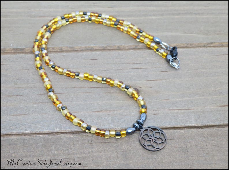 May include: A beaded necklace featuring a circular pendant with a geometric flower-like cutout design. The necklace is composed of small, translucent yellow, amber, and metallic gray beads arranged in a repeating pattern, finished with a metal clasp.