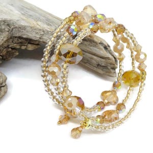 Puede incluir: Una pulsera de m&uacute;ltiples hilos con cuentas doradas y transparentes. La pulsera presenta cuentas de varios tama&ntilde;os y formas, incluidas cuentas facetadas y redondas. Las cuentas est&aacute;n ensartadas en un alambre de color dorado. La pulsera se exhibe sobre una superficie blanca con un trozo de madera flotante.