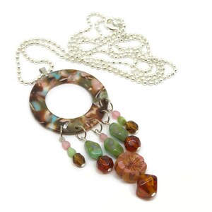 May include: A silver chain necklace with a large, round, multi-colored pendant. The pendant has a hole in the center and is decorated with a variety of colorful beads, including green, pink, and brown.