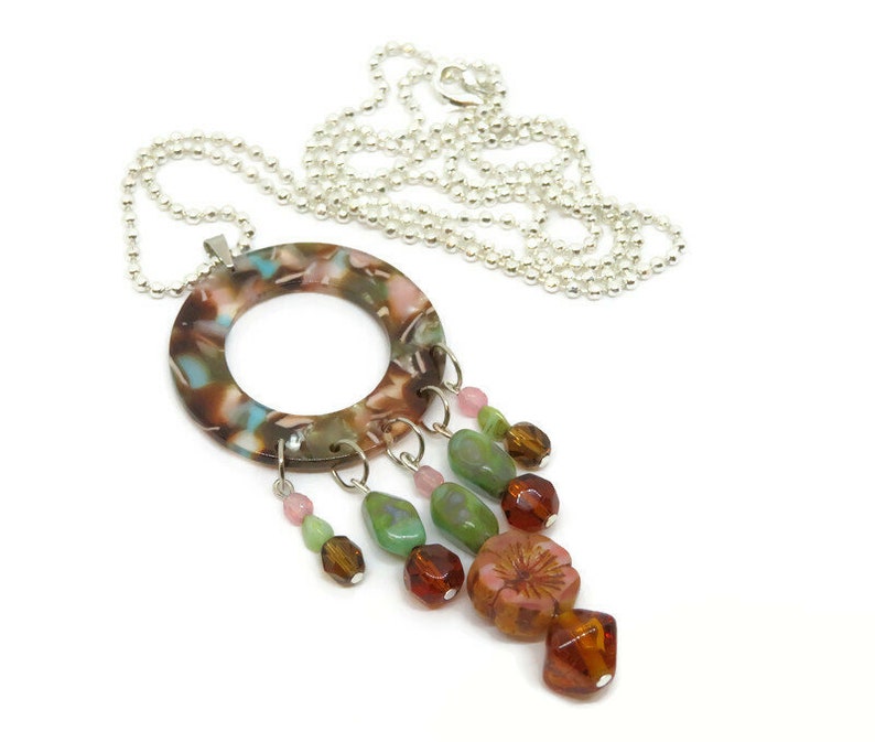 May include: A silver chain necklace with a large, round, multi-colored pendant. The pendant has a hole in the center and is decorated with a variety of colorful beads, including green, pink, and brown.