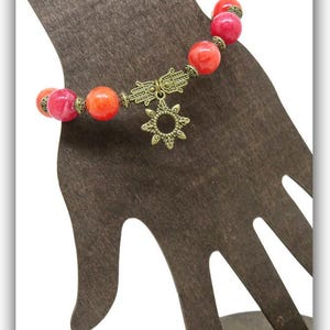 Pu&ograve; includere: Un braccialetto con perline rotonde rosse e arancioni. Il braccialetto presenta un ciondolo a mano di Hamsa color oro e un ciondolo a forma di sole. Il braccialetto &egrave; esposto su un supporto marrone a forma di mano. Il nome del negozio Etsy "MyCreativeSideJewels.etsy.com" &egrave; in basso.