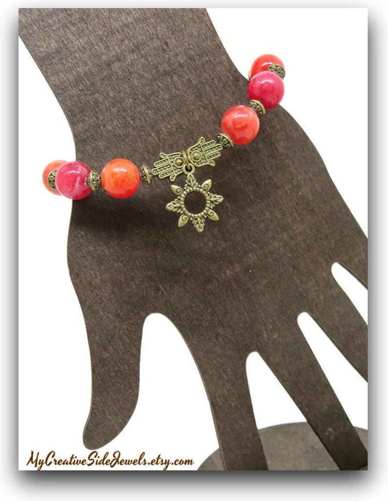 Pu&ograve; includere: Un braccialetto con perline rotonde rosse e arancioni. Il braccialetto presenta un ciondolo a mano di Hamsa color oro e un ciondolo a forma di sole. Il braccialetto &egrave; esposto su un supporto marrone a forma di mano. Il nome del negozio Etsy "MyCreativeSideJewels.etsy.com" &egrave; in basso.